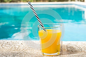 Glass of freshly pressed tropical fruits orange juice with striped straw standing on deck of swimming pool. Bright sunlight