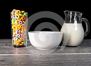 A glass full of colorful sugary cereals with a milk jug and a white ceramic bowl