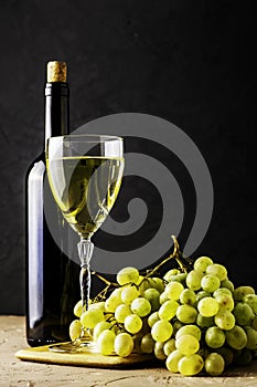 a glass glass and a bottle of white wine and a brush of white grapes on a cutting board on a black background