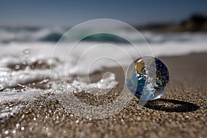 A Glass Globe Representing Earth Resting on a Sandy Beach with Ocean Waves Approaching.