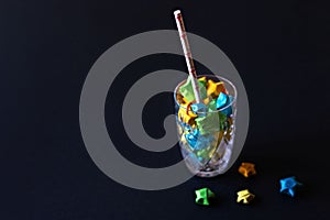 Glass goblet with paper straw and colorful origami stars on a black background. Eco-friendly decor and holiday concept