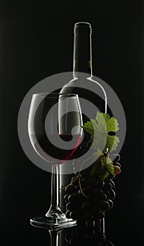 Glass goblet with red wine next to a black glass wine bottle and a bunch of grapes on a black background