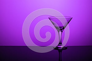 Glass goblet without wine on a thin leg stands on a mirror surface. Back light. Purple background. Copy space