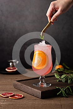 Glass of grapefruit cocktail with an orange slice in it and a person putting a mint leaf in it