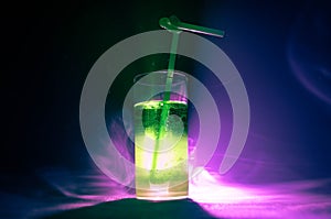 Glass of green cocktail with straw on dark background with smoke and backlights.