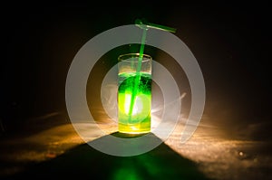 Glass of green cocktail with straw on dark background with smoke and backlights.