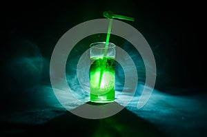 Glass of green cocktail with straw on dark background with smoke and backlights.