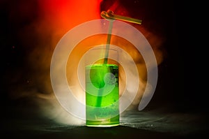 Glass of green cocktail with straw on dark background with smoke and backlights.