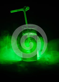 Glass of green cocktail with straw on dark background with smoke and backlights.