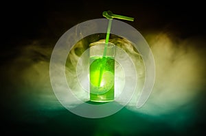 Glass of green cocktail with straw on dark background with smoke and backlights.