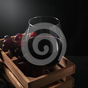 Glass half filled with red wine inside a wooden box with grapes, black background