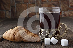 A glass of hot tea in a retro cup holder, three slices of sugar and a fresh bun on an old table. Retro stylized photo. Close-up.