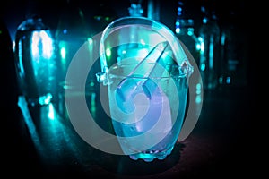 Glass ice bucket on blurred alcohol bottles background with lights and smoke. Club drinks concept. Club bar desk. Ready to serve.