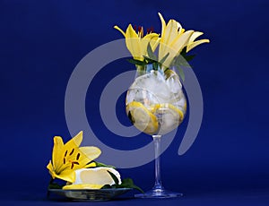 Glass with ice, cold water, lemon and fresh yellow lilies near small plate with ice cream on blue cobalt background