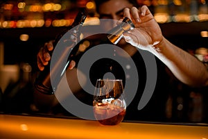 glass with ice and drink on bar and hand of bartender adds ingredient from jigger