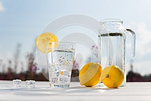 Glass of ice and water decorated with a slice of lemon standing on a white table against a decanter with water and two