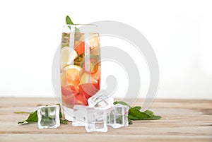 A glass of iced lemon mixed fruit tea with fresh mint leaves and ice cubes scattered on the table