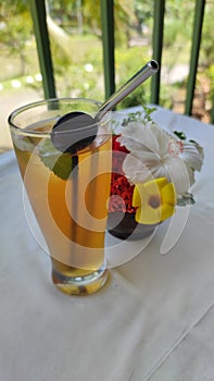 A Glass Of Iced Lemon Tea With Stainless Straw Beside A Pot Of Fresh Flowers On White-Clothed Table