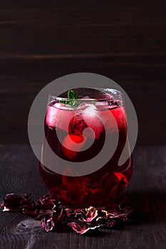 Glass of iced red hibiscus tea and dry flowers on a wooden table.