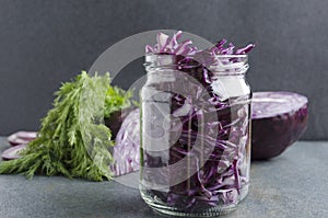 Glass jar with chopped fresh red cabbage,dill and rings of onion- preparation for fermentation on the grey surface in the kitchen.