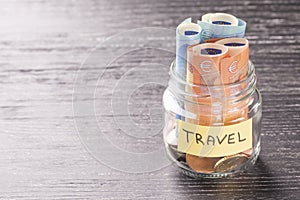 Glass jar with coins and euro banknotes with the word TRAVEL on a black wooden table
