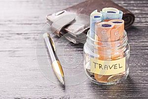 Glass jar with coins and euro notes with the word TRAVEL on a black wooden table. Leather wallet and pen