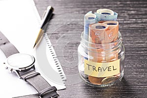 Glass jar with coins and euro notes with the word TRAVEL on a black wooden table. Wrist watch, pen and blank paper