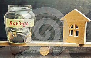 A glass jar with coins and the inscription `Savings` and house are on the scales. The concept of real estate purchase. Buy a house