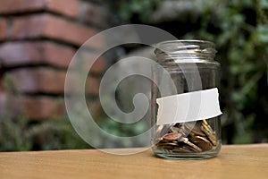 Glass jar with coins inside and with a label with a wooden background, plants and bricks.
