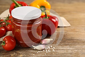 Glass jar of delicious canned lecho and fresh ingredients on wooden table, closeup. Space for text