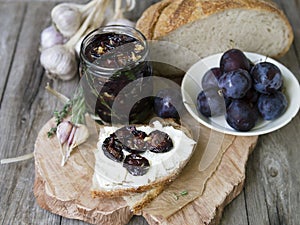 Glass jar with dried plums and fresh rosemary on wooden serving board, selective focus. Sandwich with gray bread, cream cheese and