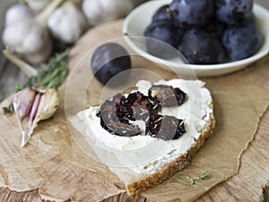 Glass jar with dried plums and fresh rosemary on wooden serving board, selective focus. Sandwich with gray bread, cream cheese and