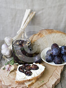 Glass jar with dried plums and fresh rosemary on wooden serving board, selective focus. Sandwich with gray bread, cream cheese and