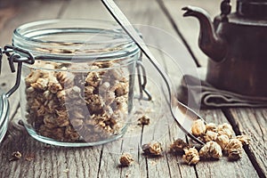 Glass jar of dry healthy chamomile buds, spoon and vintage teapot on old n wooden table.