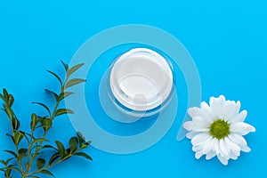 Glass jar of facial cream with white chamomile on blue background, skin care concept. Herbal lotion in bottle, natural cosmetic