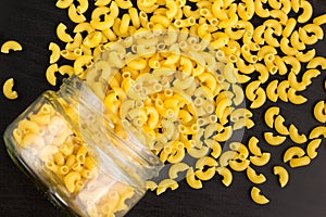 Glass jar filled with dry pasta horns and scattered around on black background