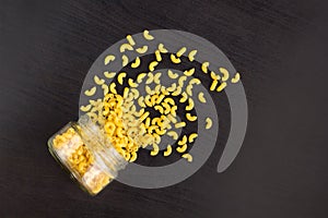 Glass jar filled with dry pasta horns and scattered around on black background