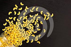 Glass jar filled with dry pasta horns and scattered around on black background