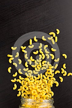 Glass jar filled with dry pasta horns and scattered around on black background