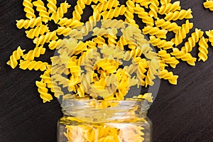 Glass jar filled with dry pasta spiral fusilli and scattered around on black background