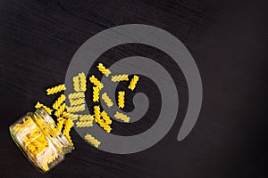 Glass jar filled with dry pasta spiral fusilli and scattered around on black background