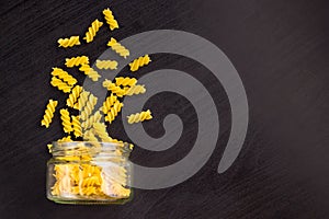 Glass jar filled with dry pasta spiral fusilli and scattered around on black background