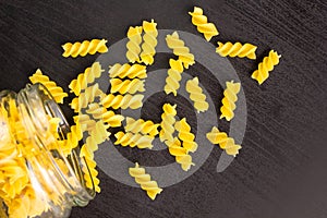 Glass jar filled with dry pasta spiral fusilli and scattered around on black background