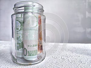 Glass Jar Filled With Euro Banknotes on a Textured White Surface. Euro currency in clear jar as background. Concept of