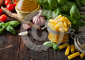 Glass jar of fresh raw penne pasta with oil and garlic, basil plant and tomatoes on wooden background
