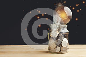 Glass jar with full of coins and light bulb on top on wooden table to show concept saving money, hope, future, education, home
