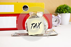 A glass jar full of coins sits on a stack of dollar bills on a work table. There is a sticker with the text TAX.