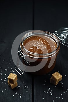 A glass jar with homemade salted caramel and a few pieces of brown sugar with salt crystals