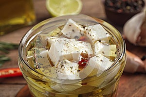 Glass jar of marinated feta cheese on wooden table, closeup