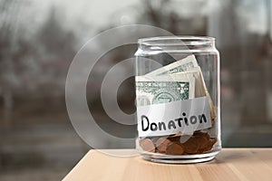 Glass jar with money and label DONATION on table against blurred background.